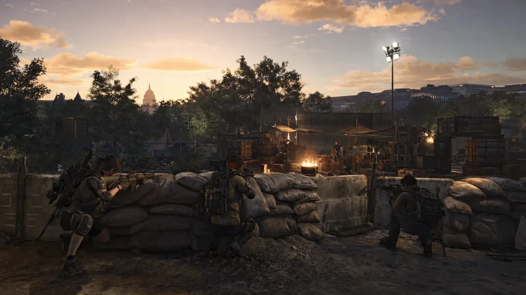 Division 2 agents defend a sandbag barricade at a fortified settlement at dusk, stacked crates of crafting materials and supplies behind the